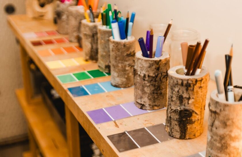 Tree trunk pencil holders with colored pens and pencils are arranged on a wooden table alongside paint color swatches sorted by hue.