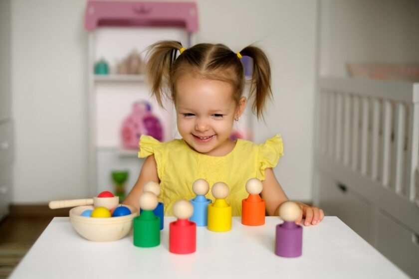 A young girl in a yellow dress plays with colorful wooden toys at a white table in a bright room, smiling and looking at the toys.