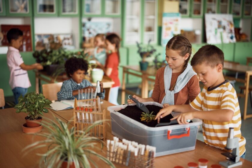 Children in a classroom work together planting a seedling in a container of soil, while other students tend to plants and work on science projects in the background.
