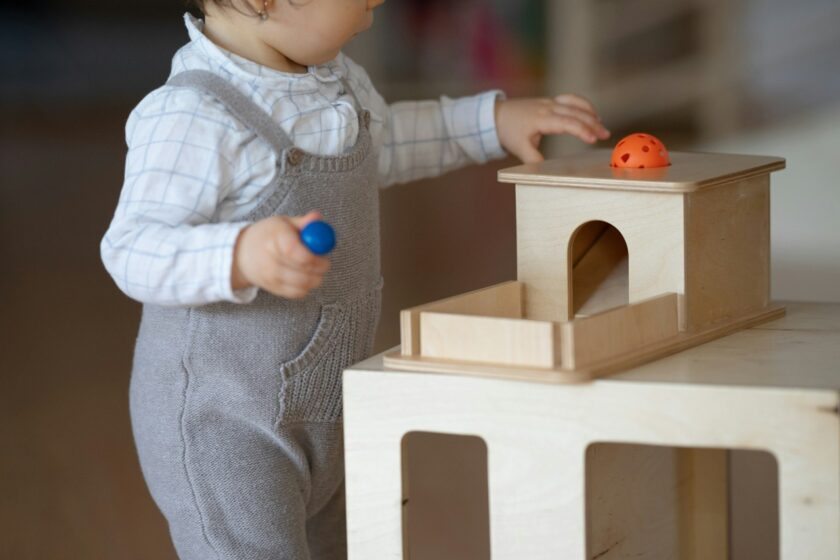 A toddler in gray overalls plays with a wooden toy, holding a blue ball and reaching toward an orange ball on top of a box.