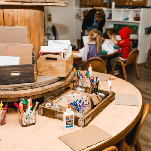 A classroom art station with supplies such as markers, scissors, and glue on a round table; children and an adult work together at another table in the background.