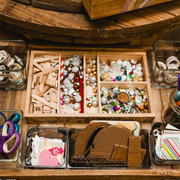A wooden tray holds jars of scissors, pens, markers, washi tape, wooden letters, beads, sequins, cardboard pieces, and patterned paper for crafting.