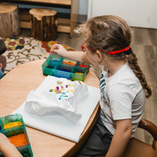 A child with braided hair and red glasses paints on a sheet of paper at a table using a tray with various paint colors.
