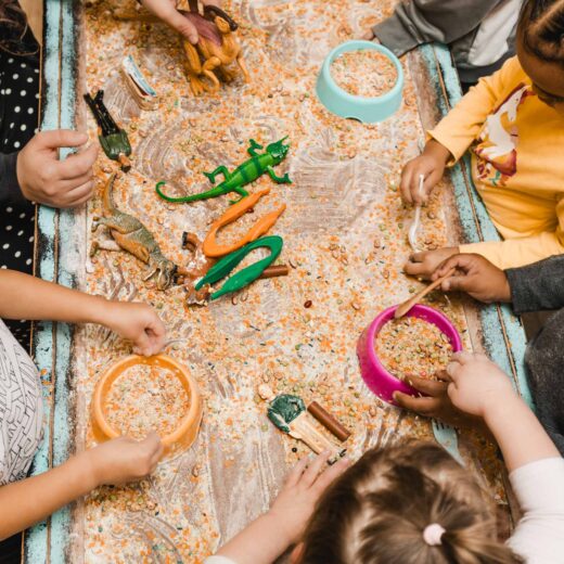 Four children play around a sensory table filled with rice, plastic dinosaurs, and bowls, using scoops and their hands to interact with the items.