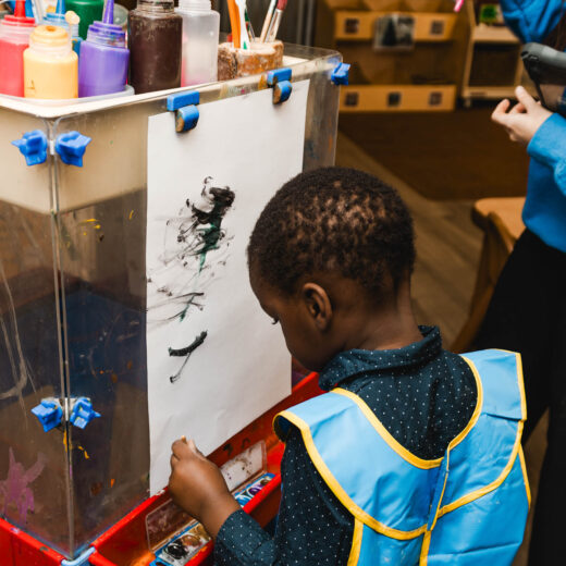 A child wearing a blue smock paints on white paper at an easel, surrounded by art supplies in a classroom setting.
