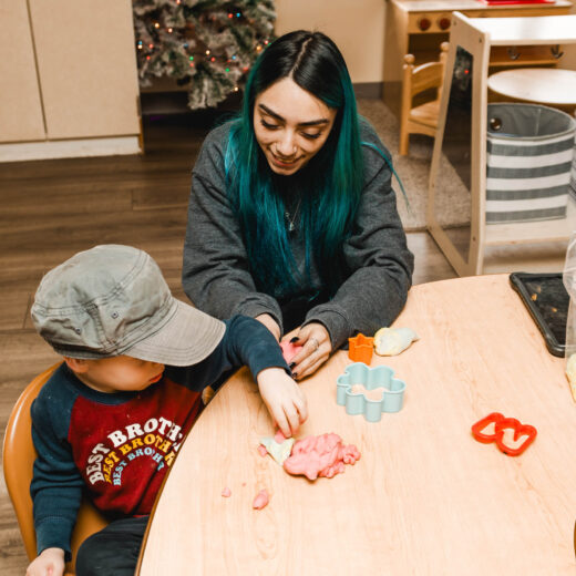 An adult and a child sit at a table playing with colorful playdough and plastic cutters in a classroom setting.
