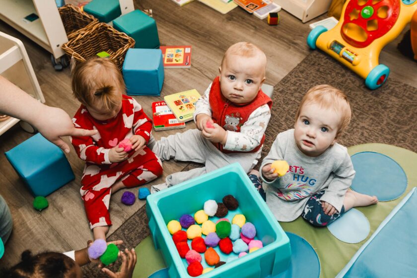 Three babies sit on the floor around a blue bin filled with colorful pom-poms, playing and looking up. Children's toys and books are visible in the background.