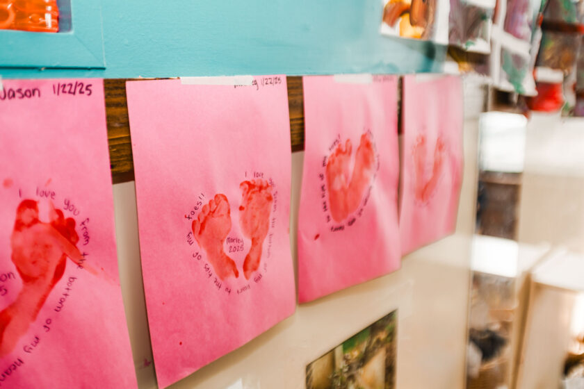 Four pink papers with red heart-shaped handprints and text are taped to a wall, likely as a children's craft or classroom display.