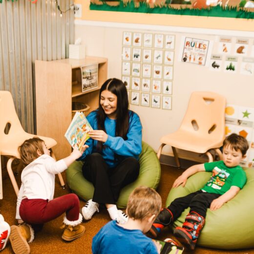 A woman reads a book to three young children in a classroom; two kids sit on bean bags, and one reaches toward the book. Educational posters decorate the walls.