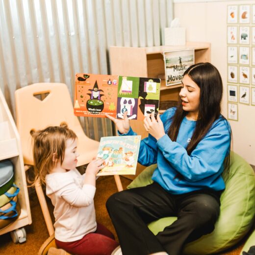 An adult and a young child sit on the floor in a classroom, reading picture books together. The adult holds up a book while the child holds another book and looks up.