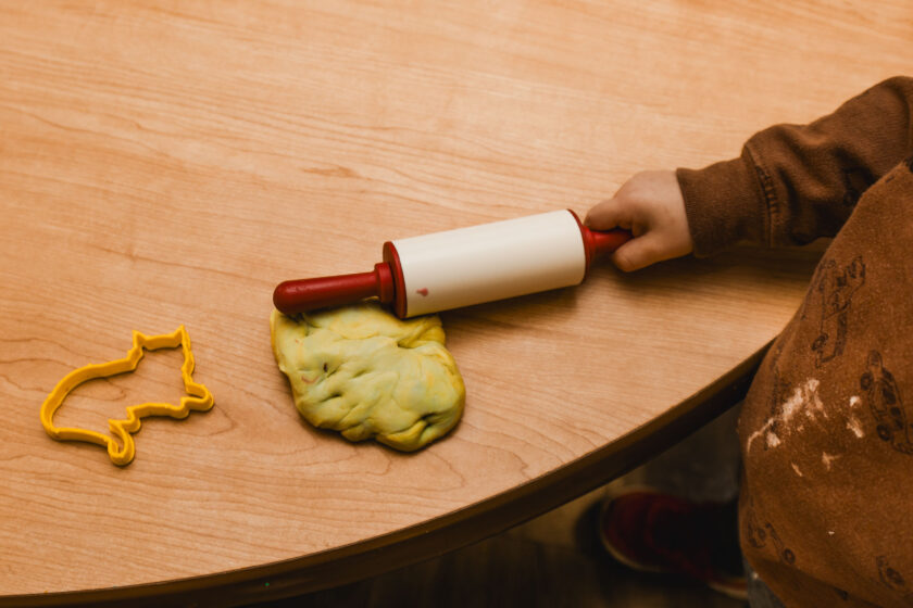A child uses a rolling pin to flatten a piece of yellow playdough on a wooden table, with a cat-shaped cookie cutter nearby.