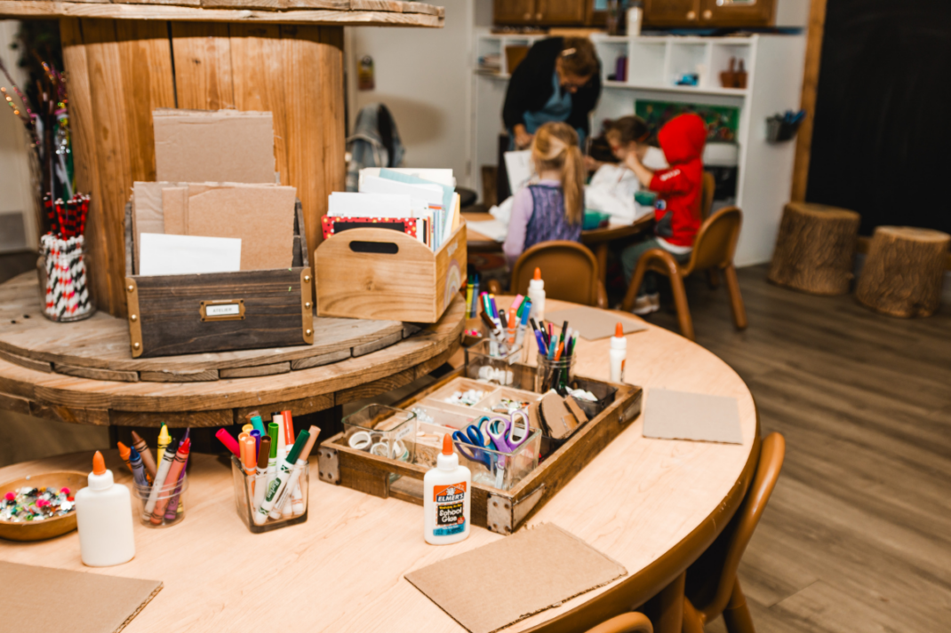 A classroom art station with markers, glue, scissors, and paper on tables; children and an adult work together at a table in the background.