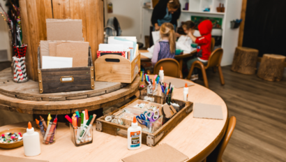 A classroom art station with markers, glue, scissors, and paper on tables; children and an adult work together at a table in the background.